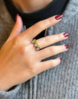 Hand with manicured nails wearing a gold ring with green and clear gemstones.
